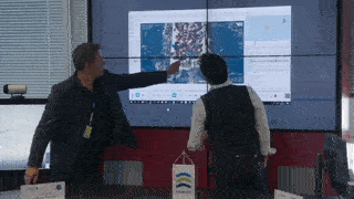 Shows two men pointing at a screen displaying a boat in the Mediterranean Sea on the AIMS-ISR mission system software.