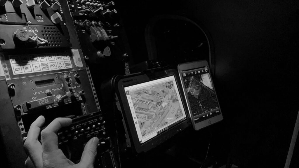 A pilot reaches for the controls beside a moving-map tablet showing mission imagery at night.
