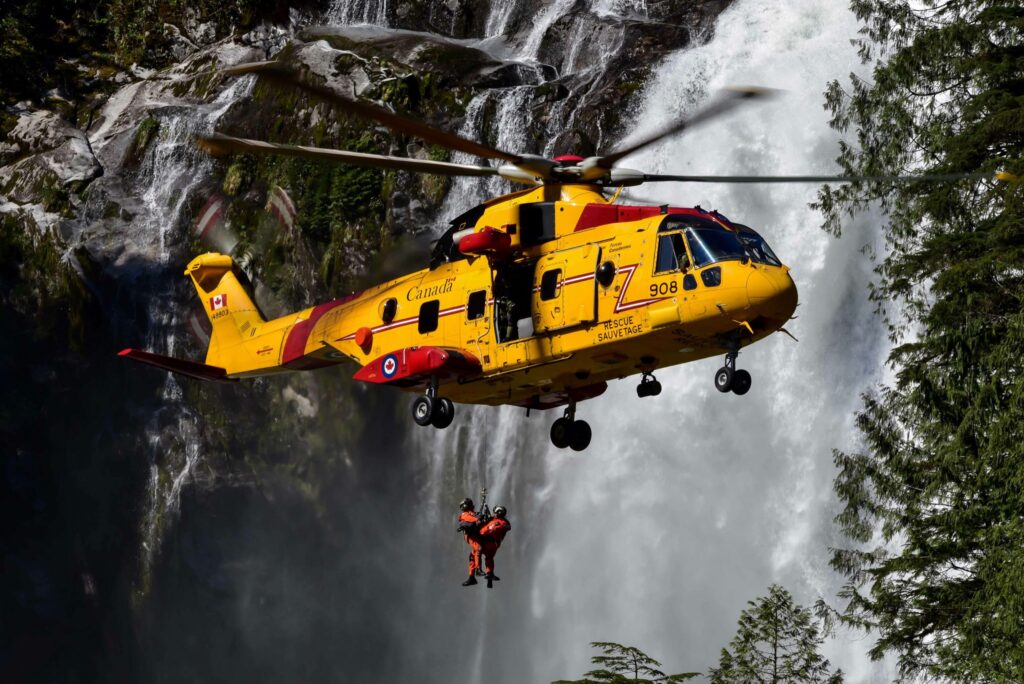A yellow search and rescue helicopter hovers beside a waterfall while hoisting a rescue technician.