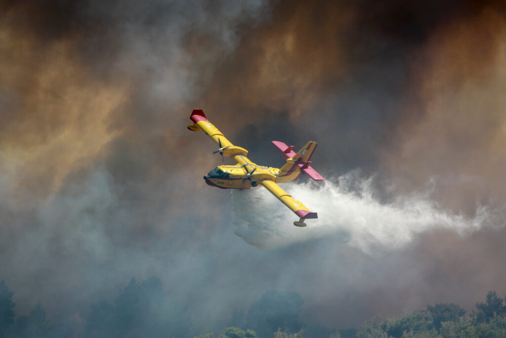 A Canadair CL-415 drops water while flying through thick wildfire smoke.