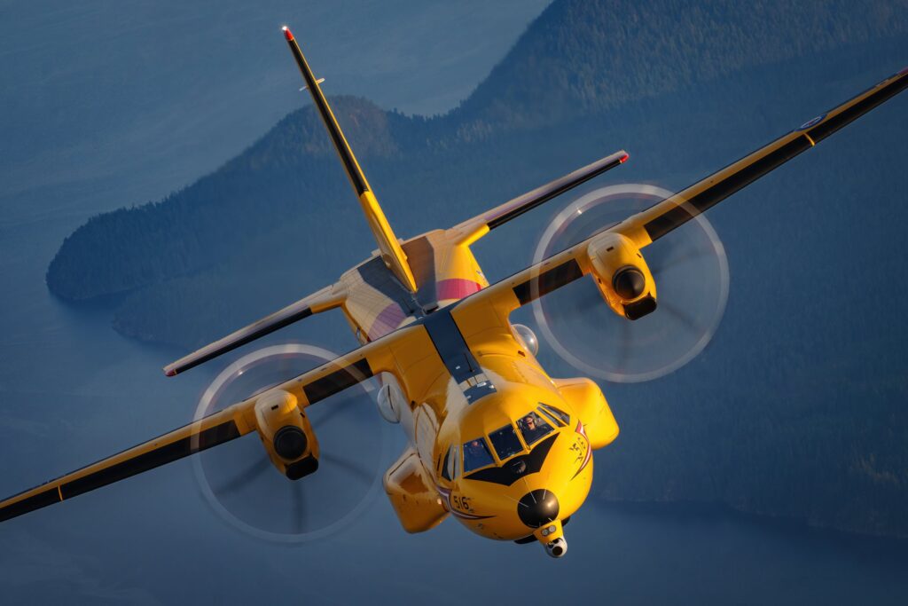 A yellow CC-295 Kingfisher search-and-rescue aircraft banks low over coastal water.