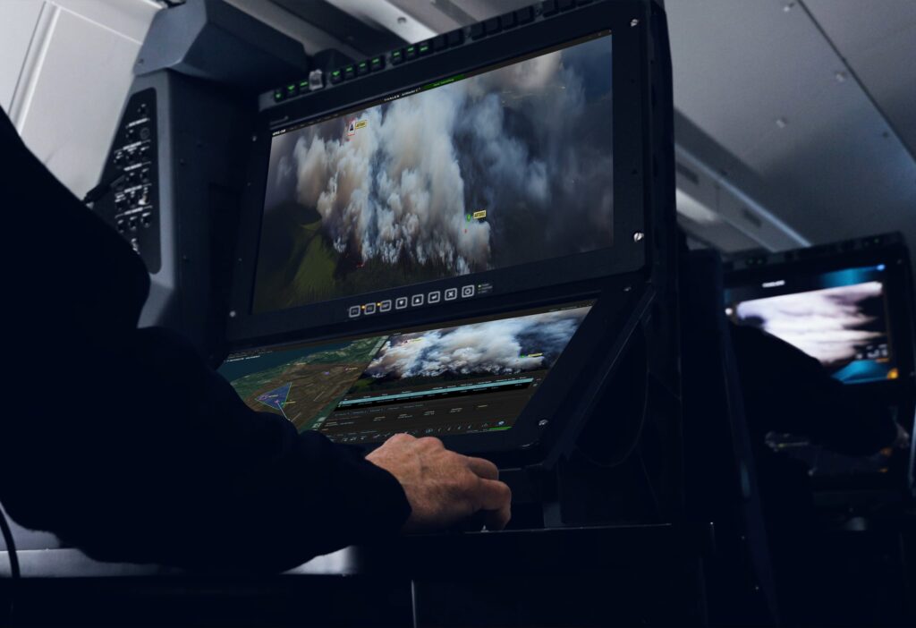 An operator works a mission console in a darkened aircraft cabin, with live sensor video on the upper display and a map on the lower one.
